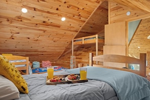 Interior de una cabaña de madera con literas, una bandeja de desayuno con fruta y jugo, y una ventana que ofrece vistas al exterior.