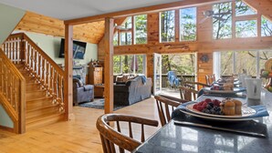 Un'ampia zona giorno con una scala in legno, un tavolo da pranzo imbandito con frutta e muffin, e ampie finestre che offrono vista all'esterno.