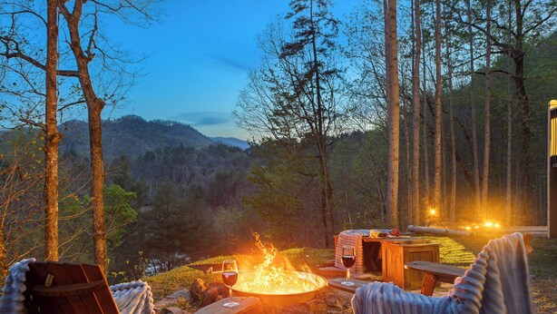Un fuoco di campo con due sedie di legno e un tavolo, circondato da alberi e una vista sulle colline.