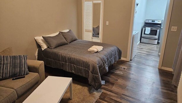 Ein Schlafzimmer mit Bett, Sofa und kleinem Tisch. Eine Tür führt zu einem Küchenbereich.