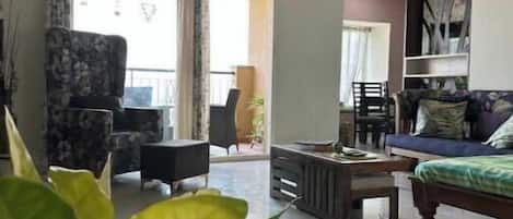 Un salon spacieux doté d’un ventilateur de plafond, d’un canapé, d’une table basse et d’un coin repas avec des chaises. On y trouve une grande fenêtre avec des rideaux, une plante en pot et une étagère fixée au mur.