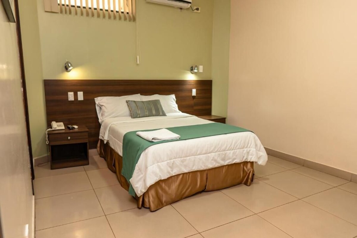 A hotel room with a bed, bedside table, and wall-mounted air conditioning unit.