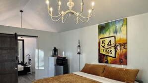 A bedroom with a large bed, a chandelier, a painting of a road sign, and a kitchenette. - Cabin 1 of 4 comfortably nestled in a country setting with amazing sunset view (Weatherford)