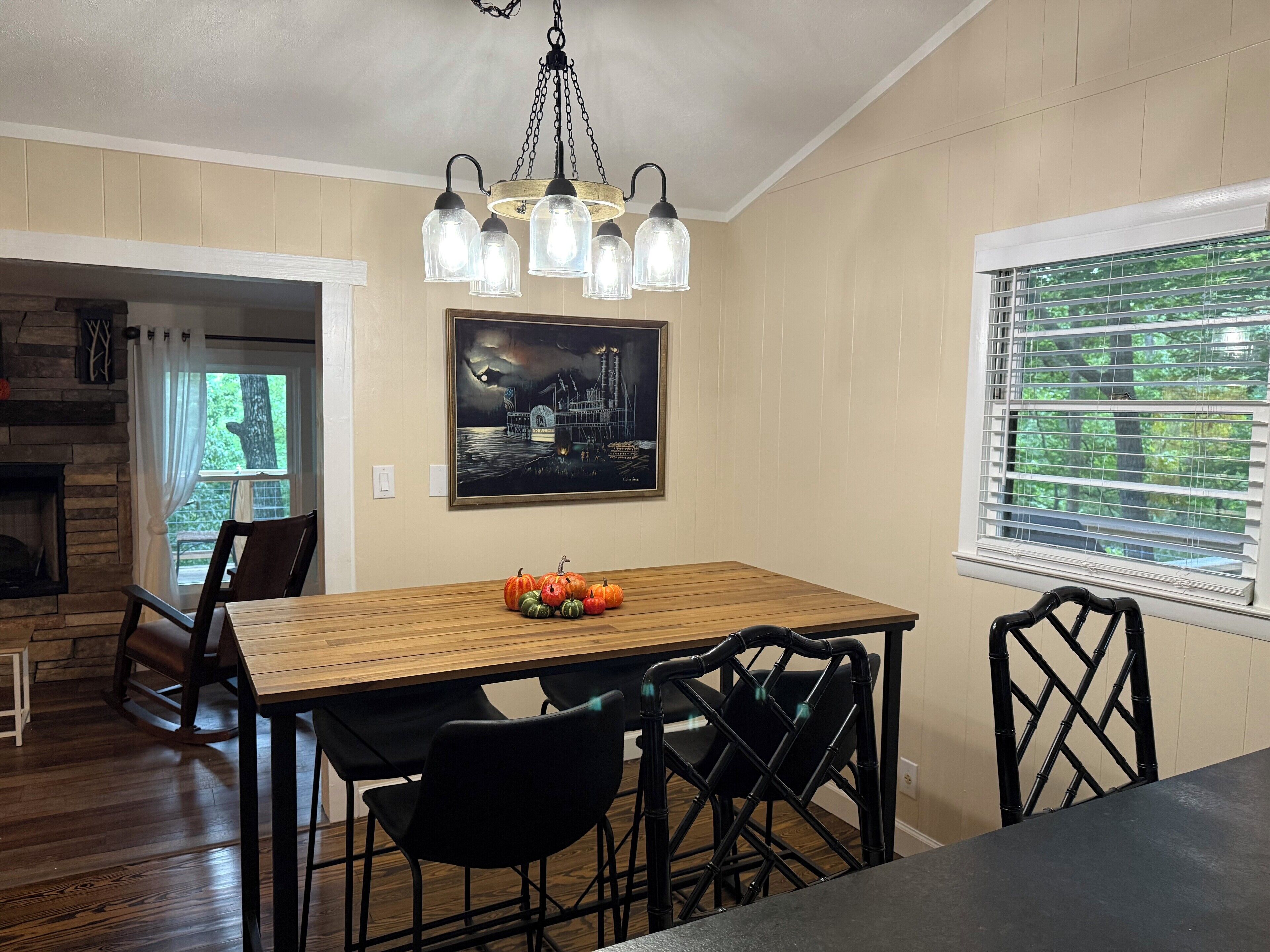 Un coin repas avec une table en bois, des chaises noires, un lustre, un tableau et une fenêtre avec des stores.