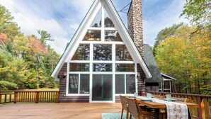 A log cabin with a large front window, a stone chimney, and a wooden deck with a table and chairs.