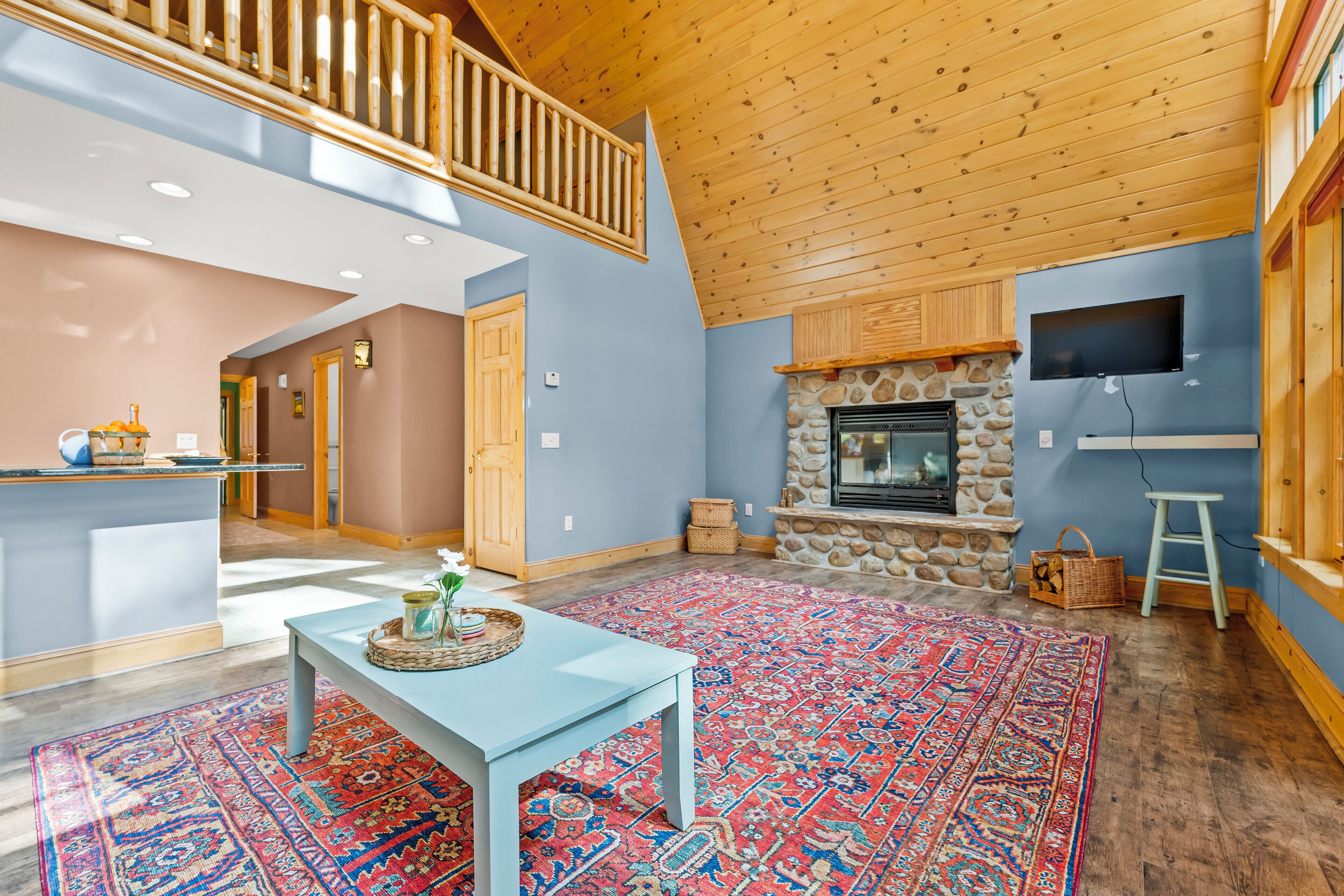 A spacious living area with a high ceiling, a stone fireplace, a television mounted on the wall, and a colorful rug.