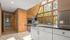 A modern kitchen with white cabinets, a stone fireplace, and large windows overlooking a forest.