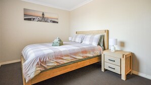 A neatly made bed with a checkered bedspread, a wooden headboard, and two bedside tables with lamps. A painting of a lighthouse is on the wall above the bed. - Pilbara Chalet Private 1x1 (Port Hedland)