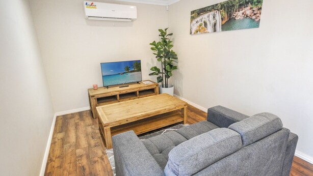 A living room with a wooden coffee table, grey sofas, a flat-screen TV, a potted plant, and a painting of a waterfall. - Pilbara Chalet Private 1x1 (Port Hedland)