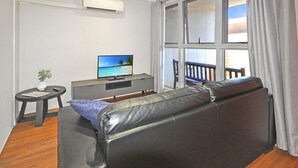 A modern living room with a leather sofa, a small side table with a plant, a grey entertainment unit with a TV displaying a beach scene, and large windows with curtains. - Substantial Studio 1x1 (Port Hedland)