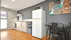 A modern kitchen with a white refrigerator, a microwave, and a coffee maker on the countertop. There is a dining table with two chairs and a painting on the wall. - Substantial Studio 1x1 (Port Hedland)
