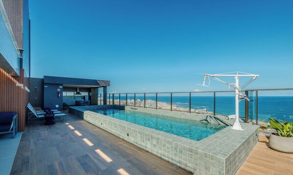 Une piscine sur le toit offrant une vue sur l’océan, un petit bar et un espace détente avec une table et des chaises.