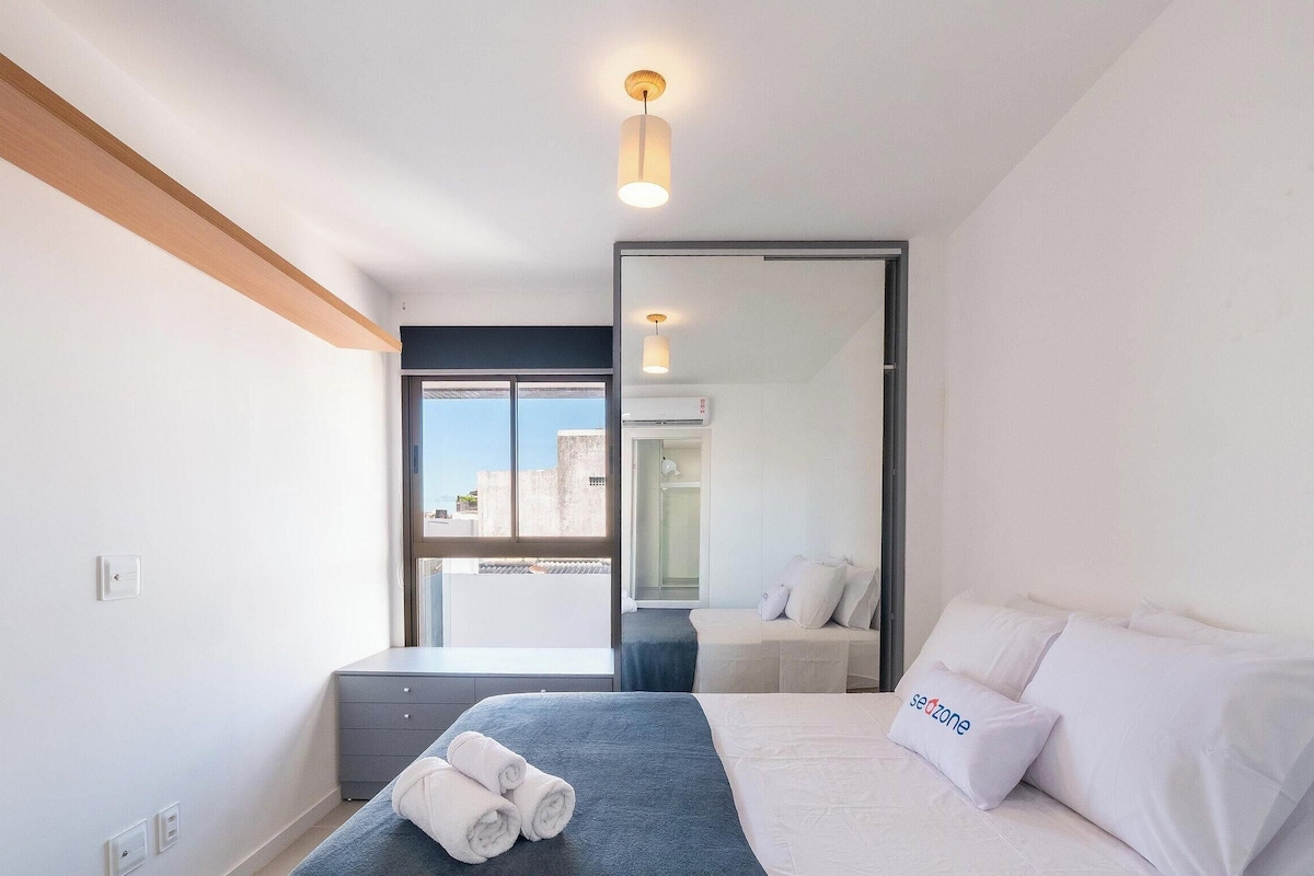 A hotel room with a bed, a small desk, a mirror, and a window with a view of buildings.