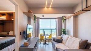 A modern living room with a TV, dining table, and yellow chairs, overlooking a balcony with a view of the sea. - Apartments in a Building With a Pool 800m From the Beach in Salvador - EDF (Salvador)