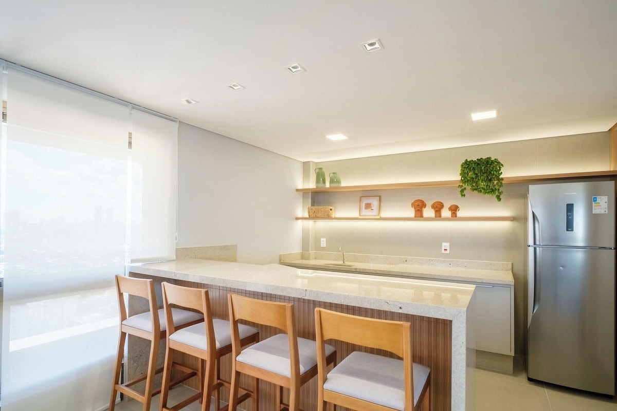 A modern kitchen with a central island, wooden chairs, a refrigerator, and a plant on the countertop.