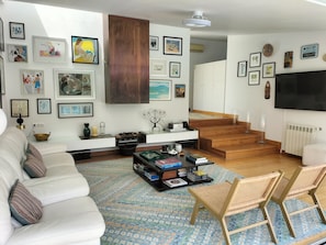 A modern living room with a white sofa, a coffee table with books, a TV, and a staircase leading to another room.