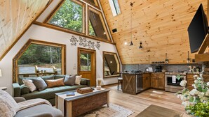 A spacious loft-style room with a large window, a cozy seating area, a kitchen with modern appliances, and a wooden ceiling. - Creekside • Sauna • Hot Tub • Fire Pit • 15 min to Town (Morganton)