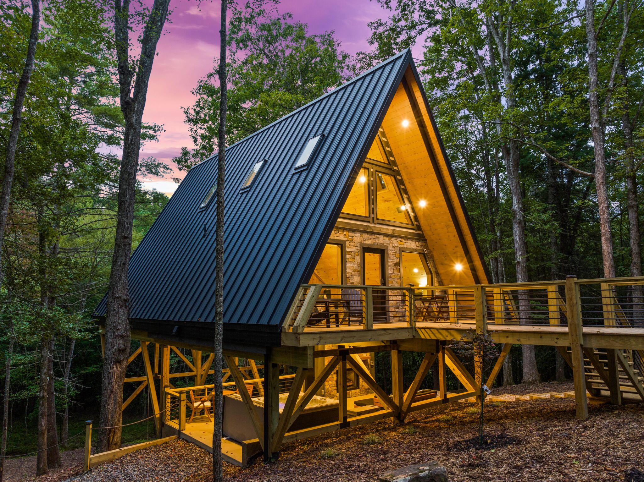 A cabin with a sloped roof, wooden deck, and stone walls, surrounded by trees.