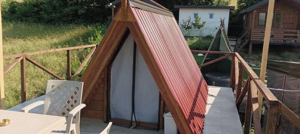 A small, triangular wooden structure with a sloped roof, possibly a shed or a small cabin, on a wooden deck with a white chair and a table.