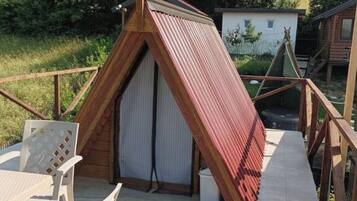 A small, triangular wooden structure with a sloped roof, possibly a shed or a small cabin, on a wooden deck with a white chair and a table.