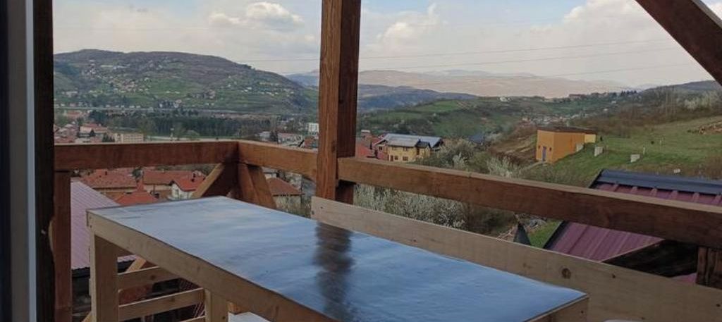 A wooden balcony with a view of a hilly landscape and buildings in the distance.