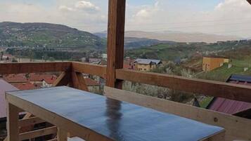 A wooden balcony with a view of a hilly landscape and buildings in the distance.