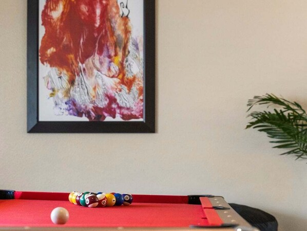 Une table de billard avec des billes et des queues, un tableau abstrait encadré accroché au mur, et une plante en pot placée sur le côté.