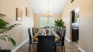 Een eetruimte met een lange tafel gedekt voor zes personen, omringd door zwarte stoelen met lederen bekleding. Een kroonluchter hangt aan het plafond, en er zijn ingelijste foto's aan de muur.