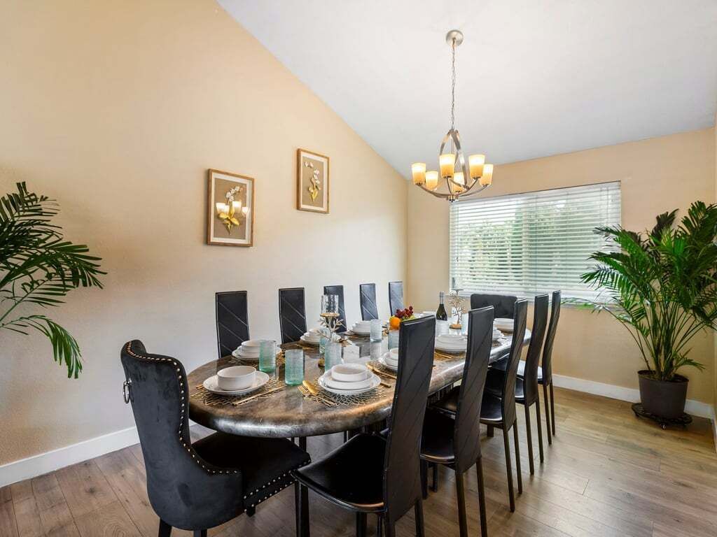 Un coin repas avec une table ronde dressée pour six personnes, entourée de chaises noires. Deux tableaux encadrés ornent le mur, un lustre est suspendu au-dessus de la table et une plante en pot se trouve à droite.