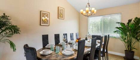 Un coin repas avec une table ronde dressée pour six personnes, entourée de chaises noires. Deux tableaux encadrés ornent le mur, un lustre est suspendu au-dessus de la table et une plante en pot se trouve à droite.