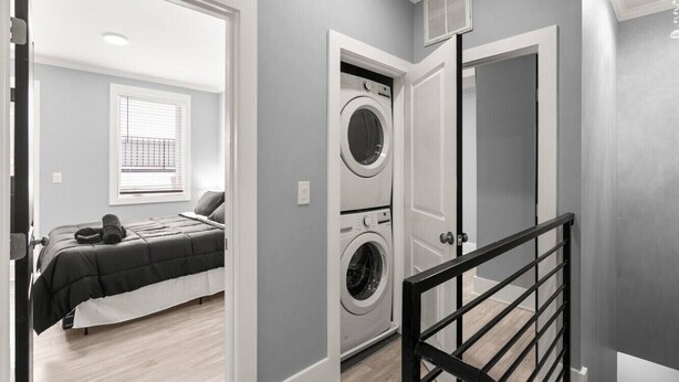 A laundry room with a washing machine and dryer, a bed with a dark-colored comforter, and a window with blinds. - DC Penthouse: Rooftop+ Views+ 3BD+ Private Parking (Washington)