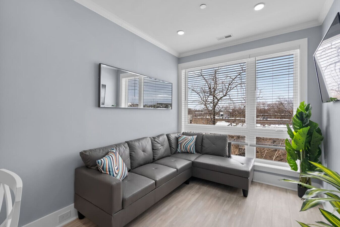 Una sala de estar moderna con un sofá modular gris, un ventanal con persianas y una planta en maceta.