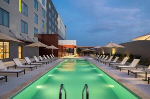Una piscina con sillas de descanso y sombrillas, rodeada por un edificio de varios pisos y ventanas.