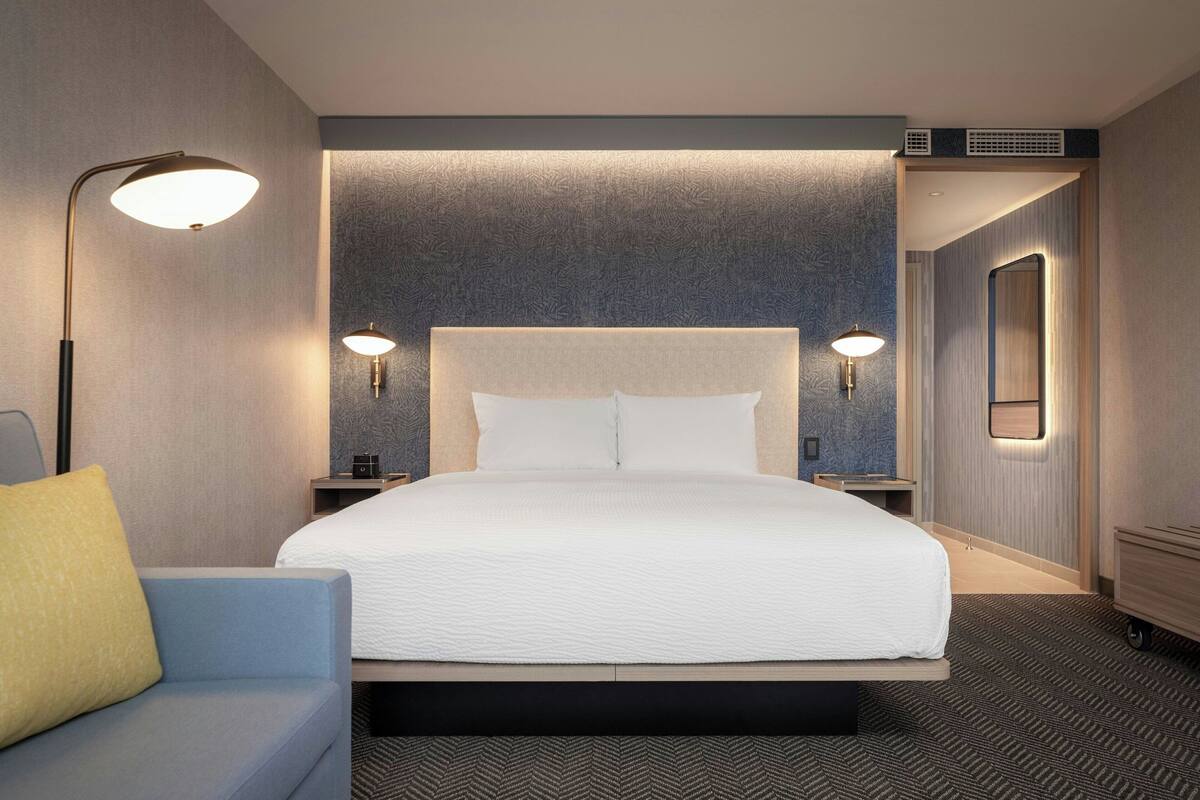Ein Hotelzimmer mit einem großen Bett, zwei Nachttischen, einem Wandspiegel und einem Sofa mit Kissen.