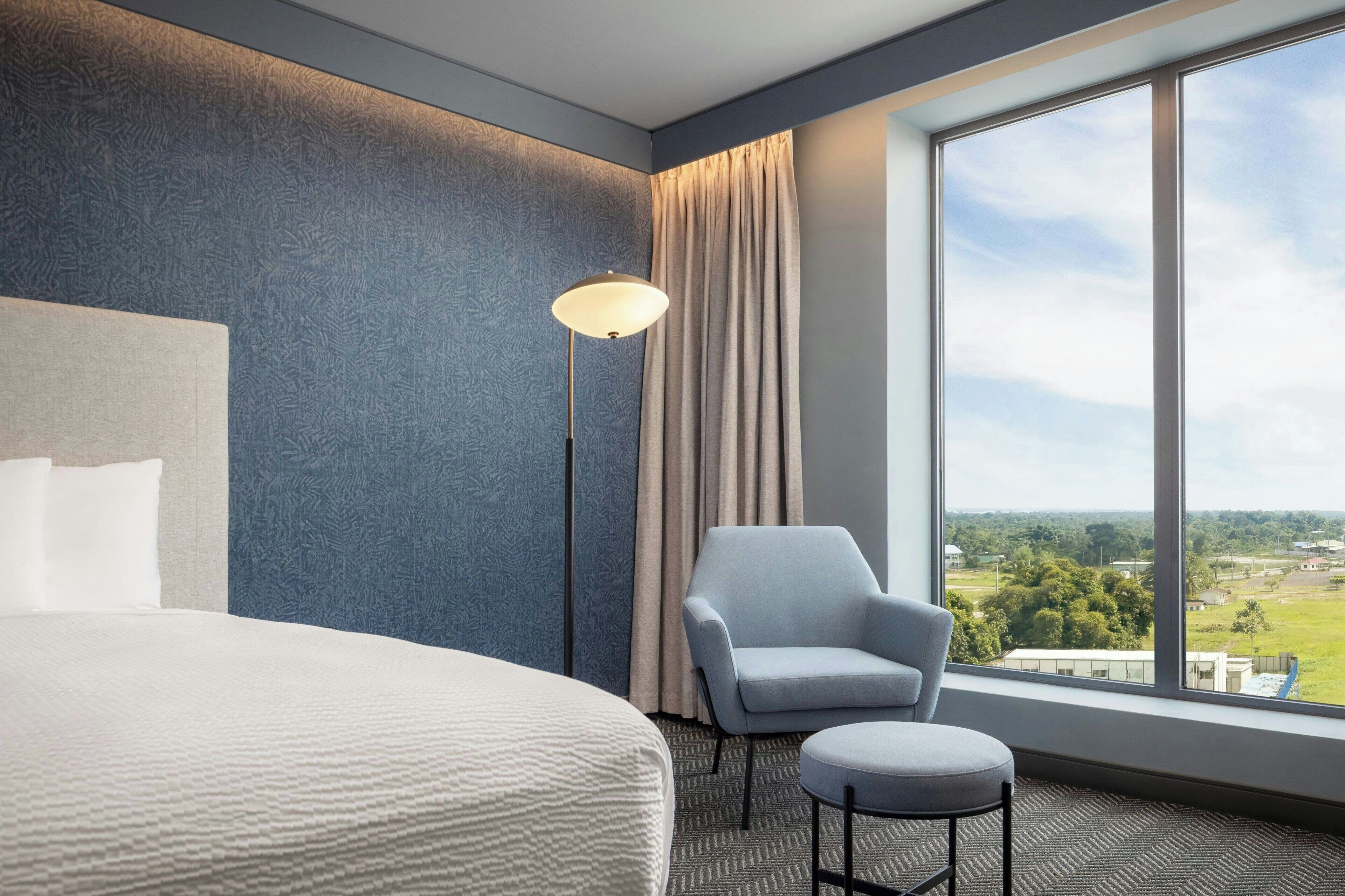A hotel room with a large bed, a chair, a small table, a floor lamp, and a large window offering a view of the outdoors.