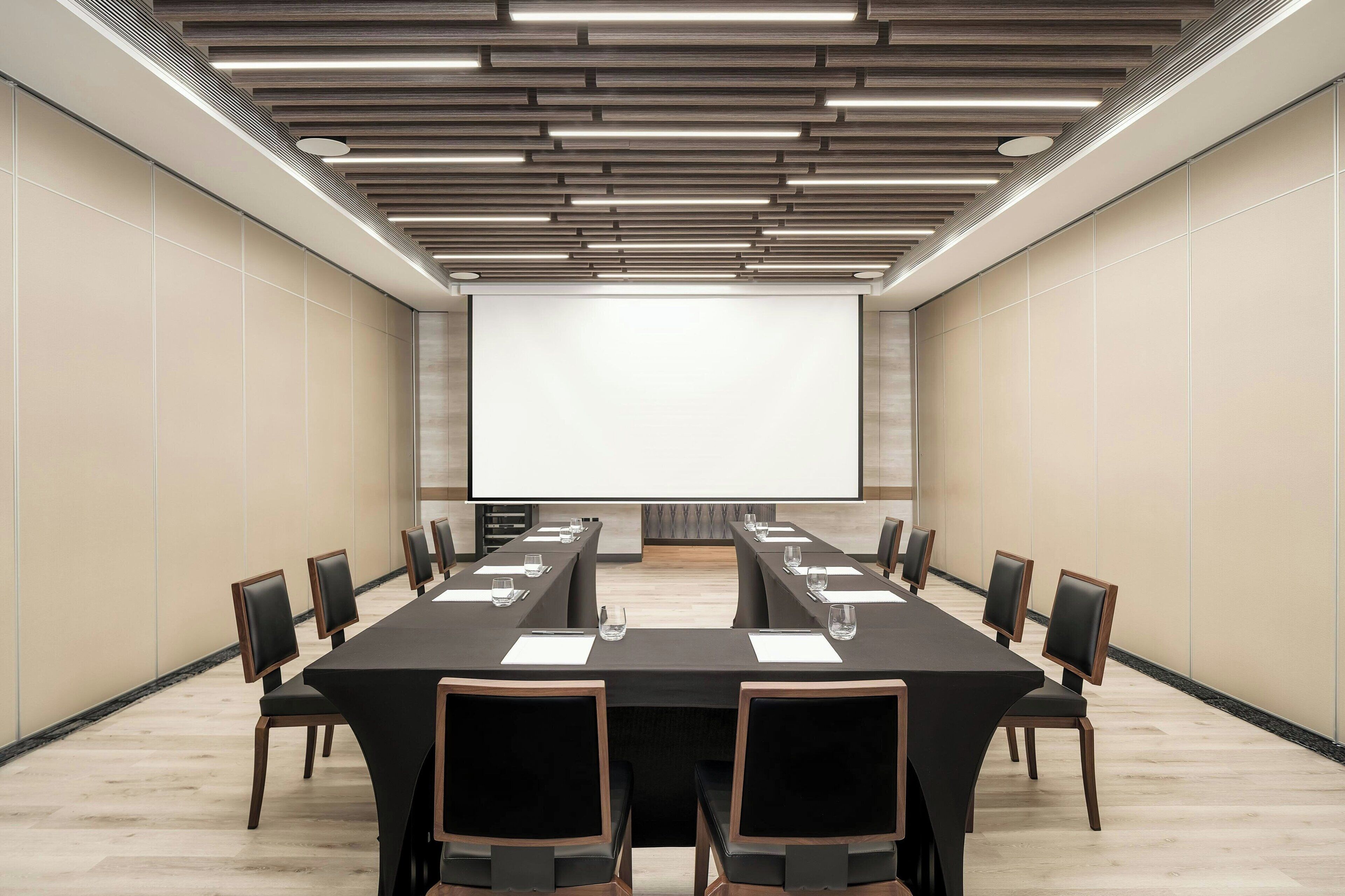 Ein Konferenzraum mit langem Tisch, Stühlen, einer Projektionsleinwand und einem modernen Deckenstil.