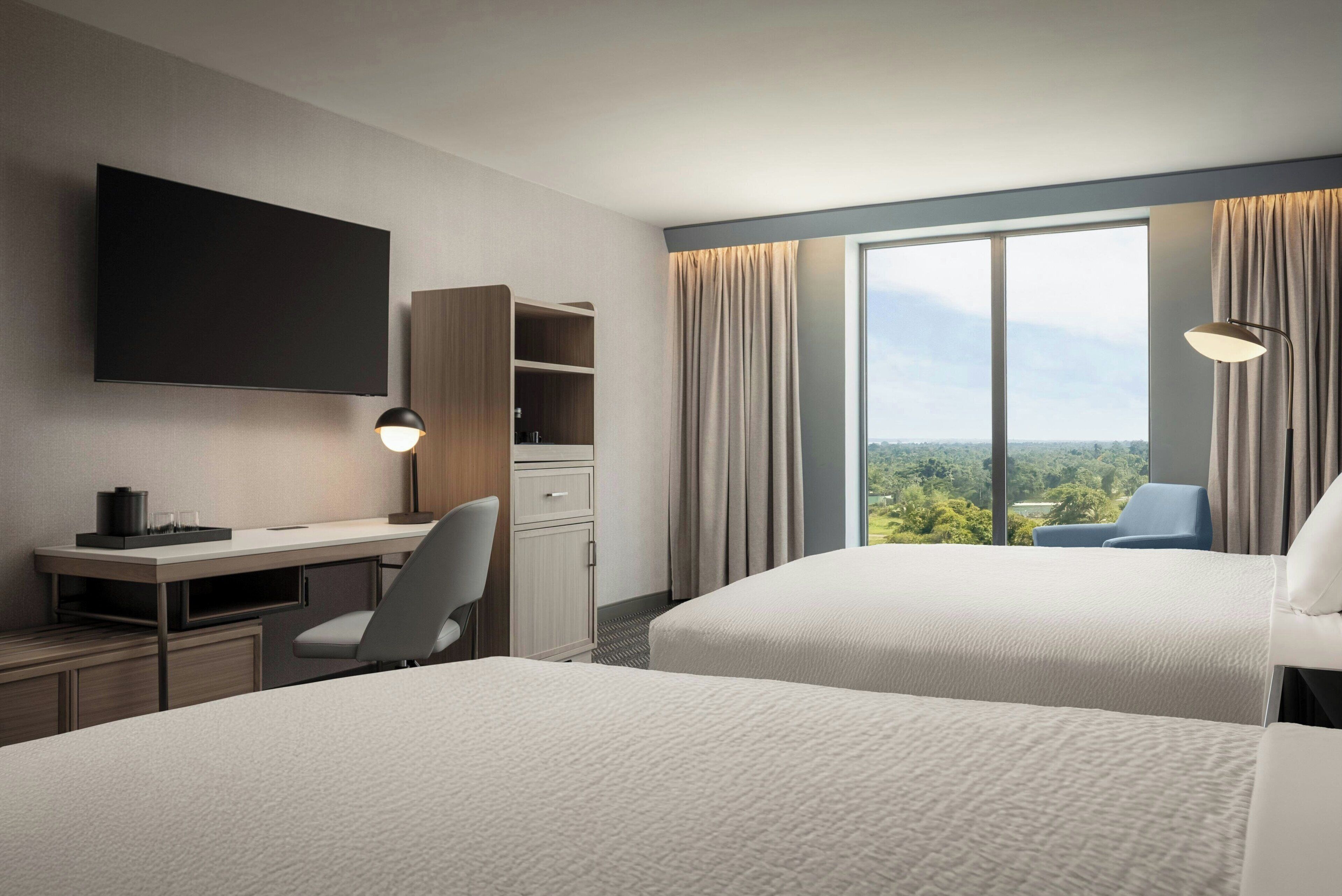 Ein Hotelzimmer mit zwei Betten, einem Schreibtisch mit Stuhl, einem Fernseher und einem großen Fenster mit Vorhängen.