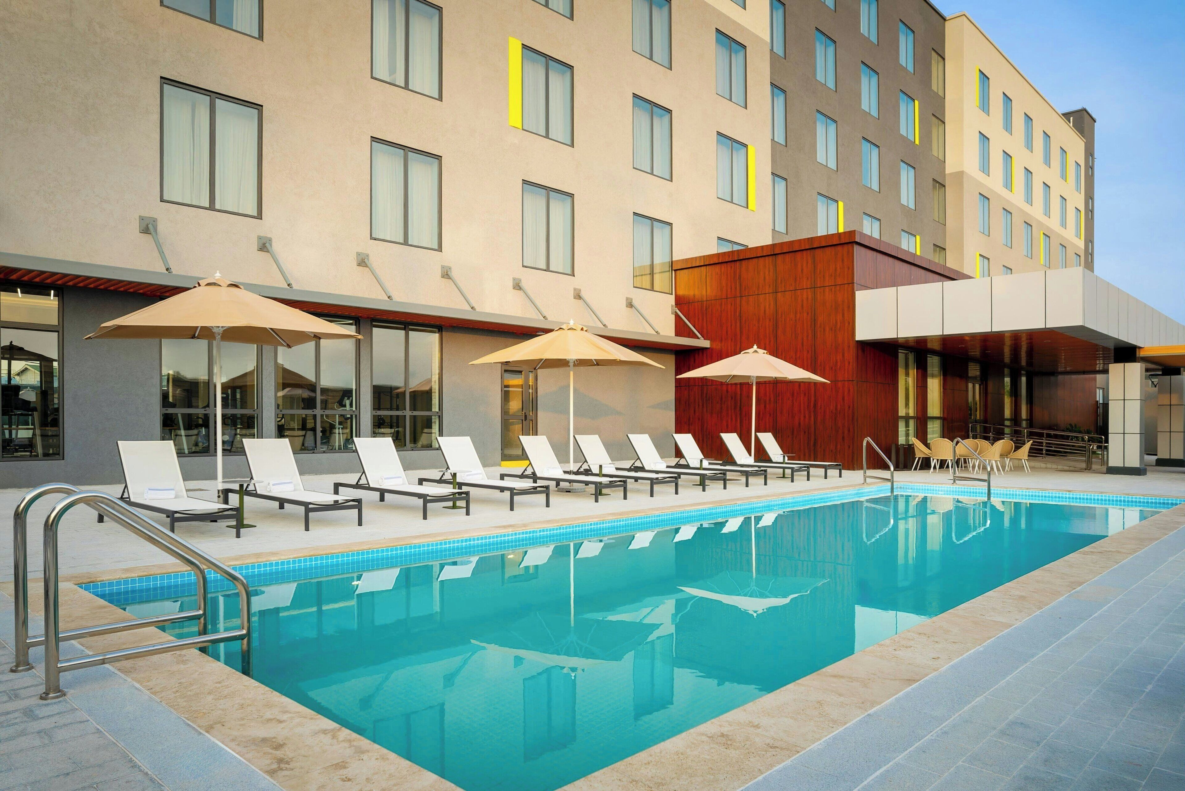 Ein Swimmingpool mit Liegestühlen und Sonnenschirmen, ein modernes Gebäude mit vielen Fenstern und einem klaren Himmel.