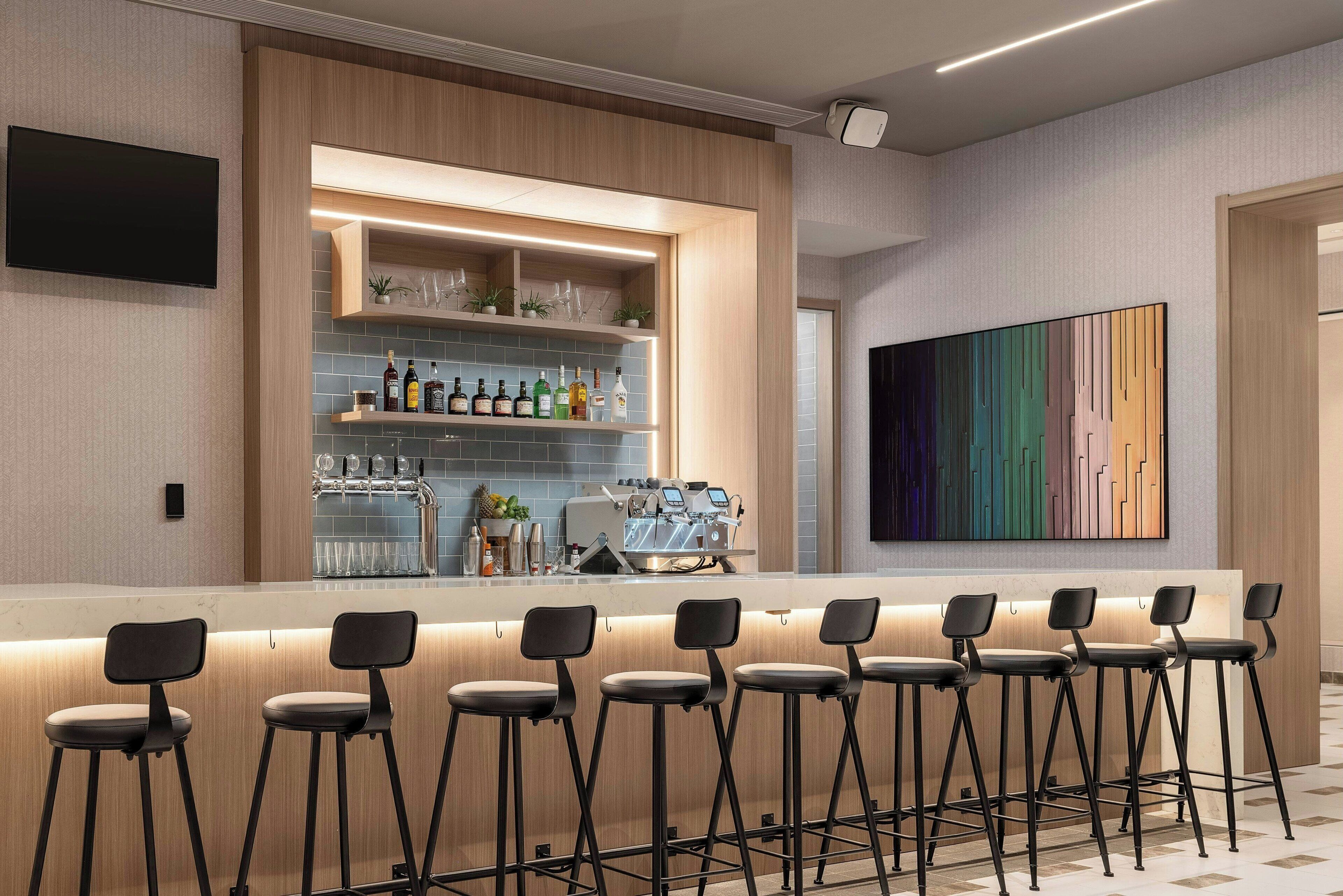 A bar area with a long counter, multiple stools, a coffee machine, and shelves with bottles and glasses.