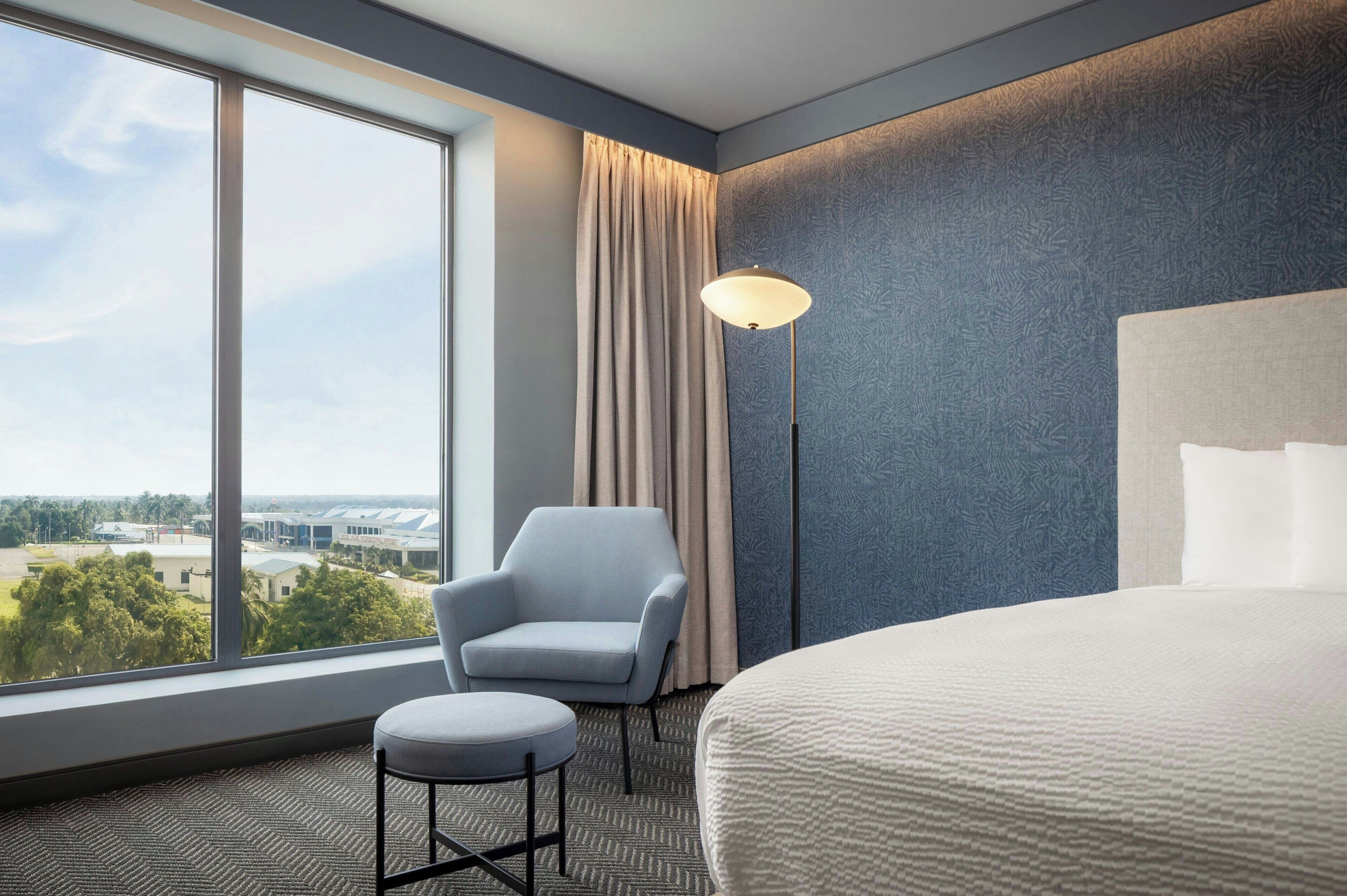 A hotel room with a large window, a bed, a chair, a small table, and a floor lamp.