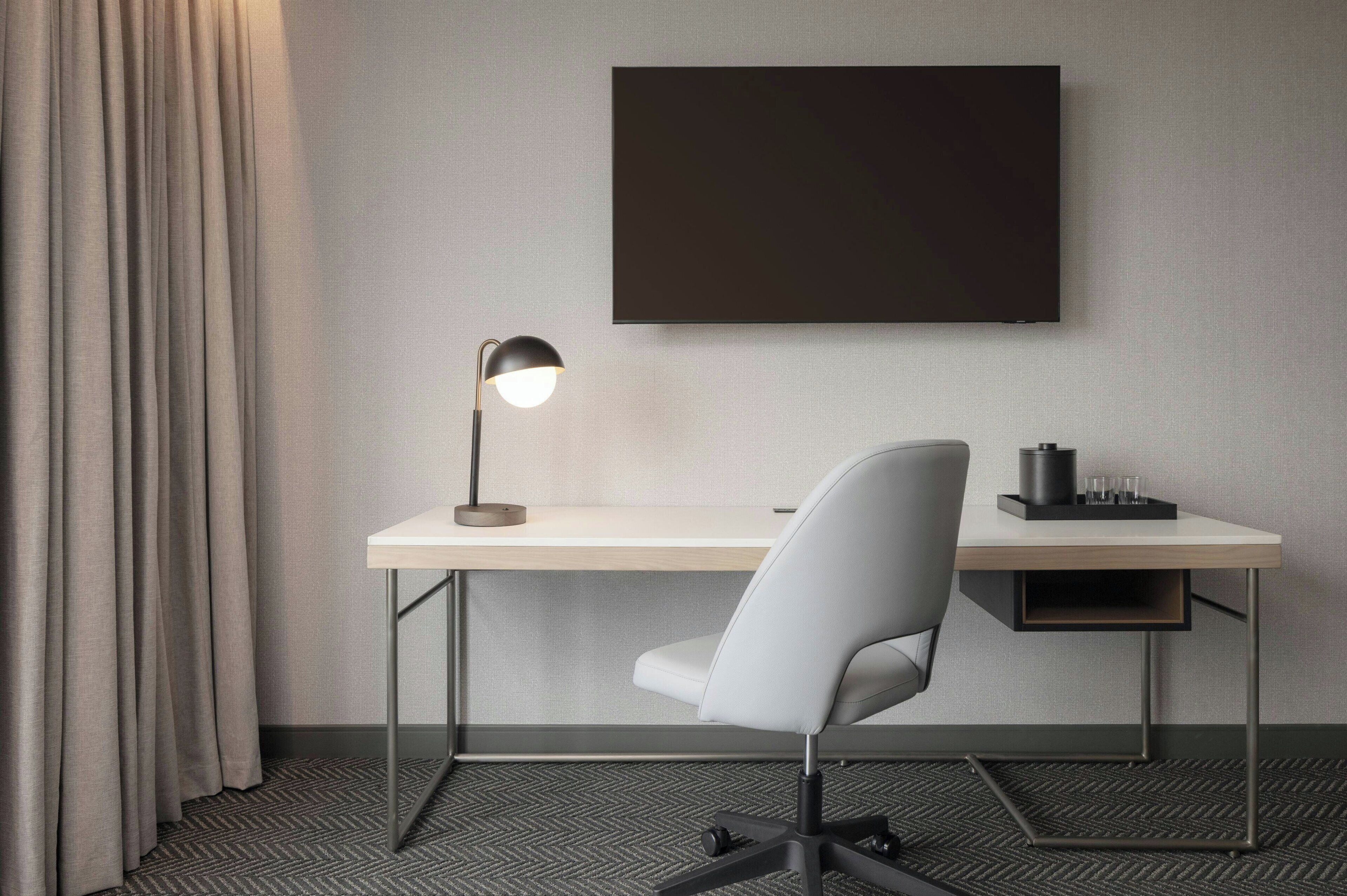 A modern hotel room with a desk, chair, lamp, and a flat-screen TV mounted on the wall.