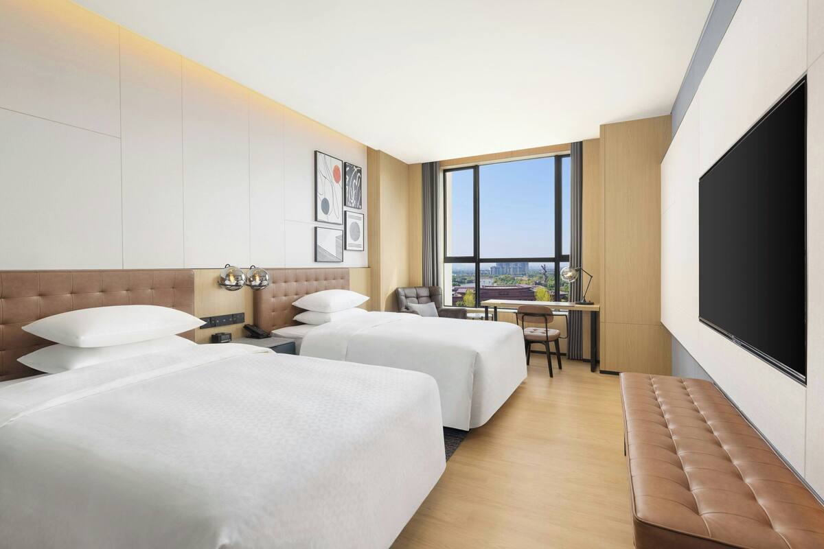 Une chambre d’hôtel avec deux lits, un bureau, une chaise, une grande télévision et une fenêtre donnant sur la ville.