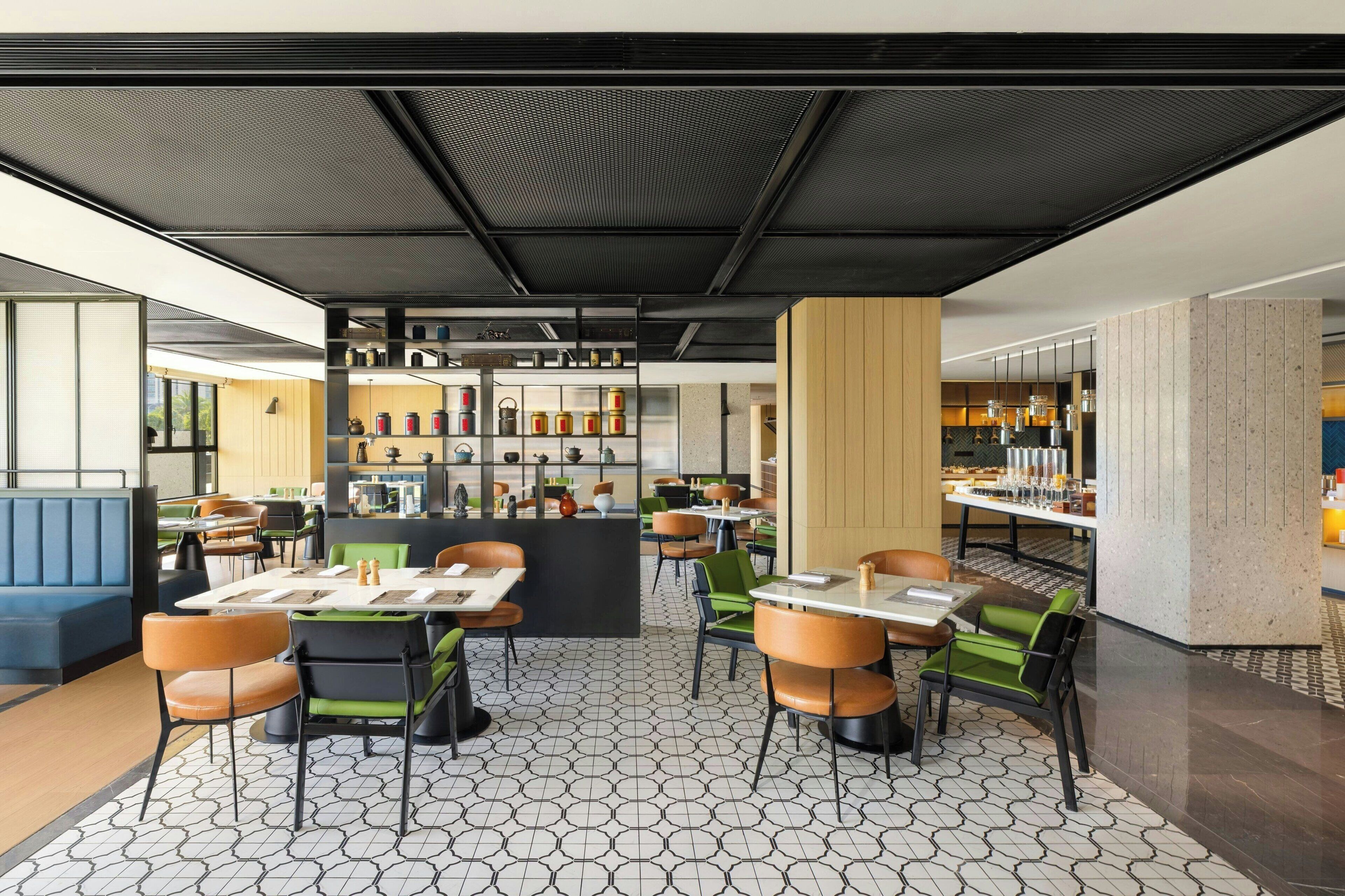 A modern restaurant interior with hexagonal patterned flooring, black and green upholstered chairs, and a central bar area with shelves of bottles and glasses.