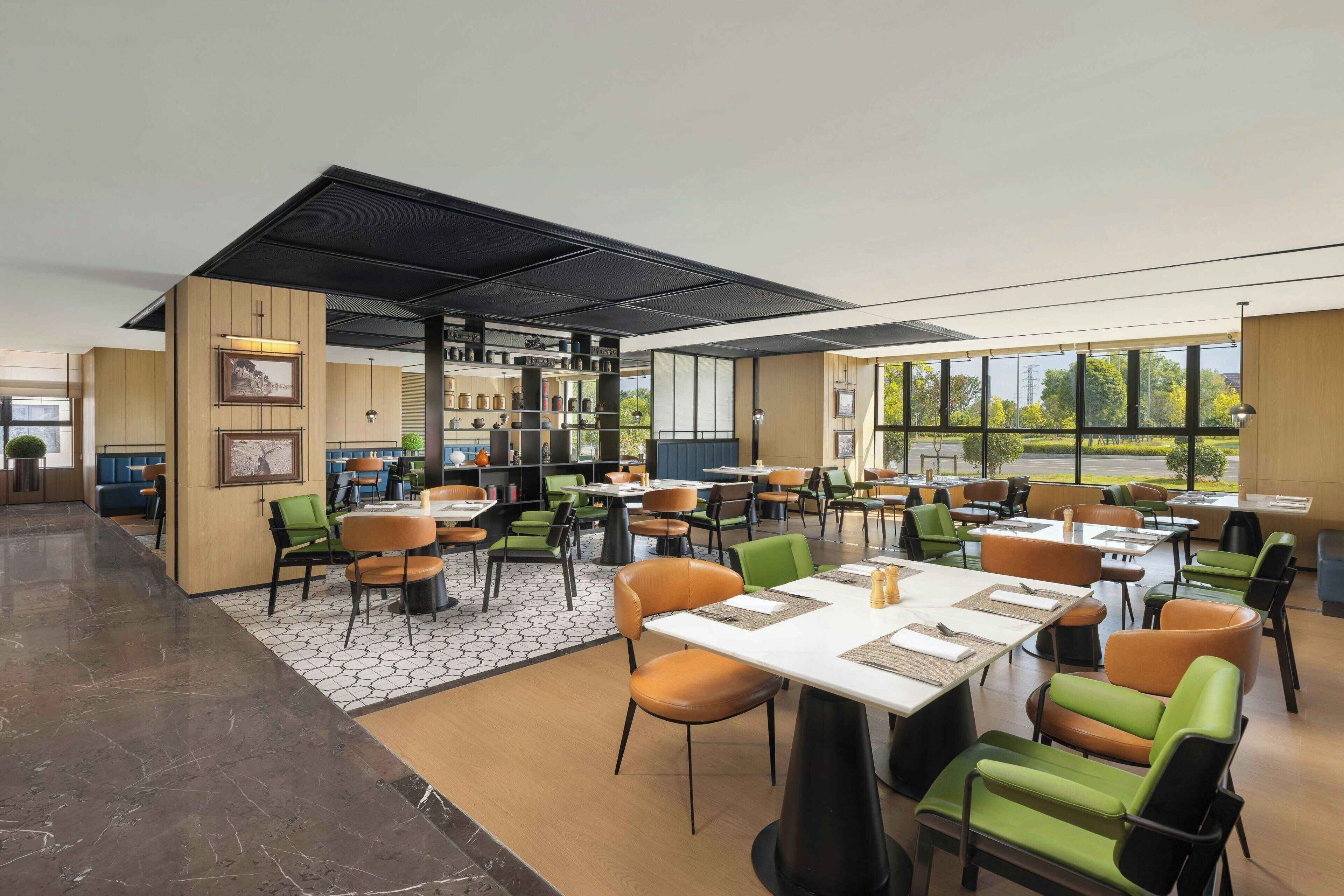 A modern restaurant with a mix of green and orange chairs, large windows, and a central open area with tables and chairs.