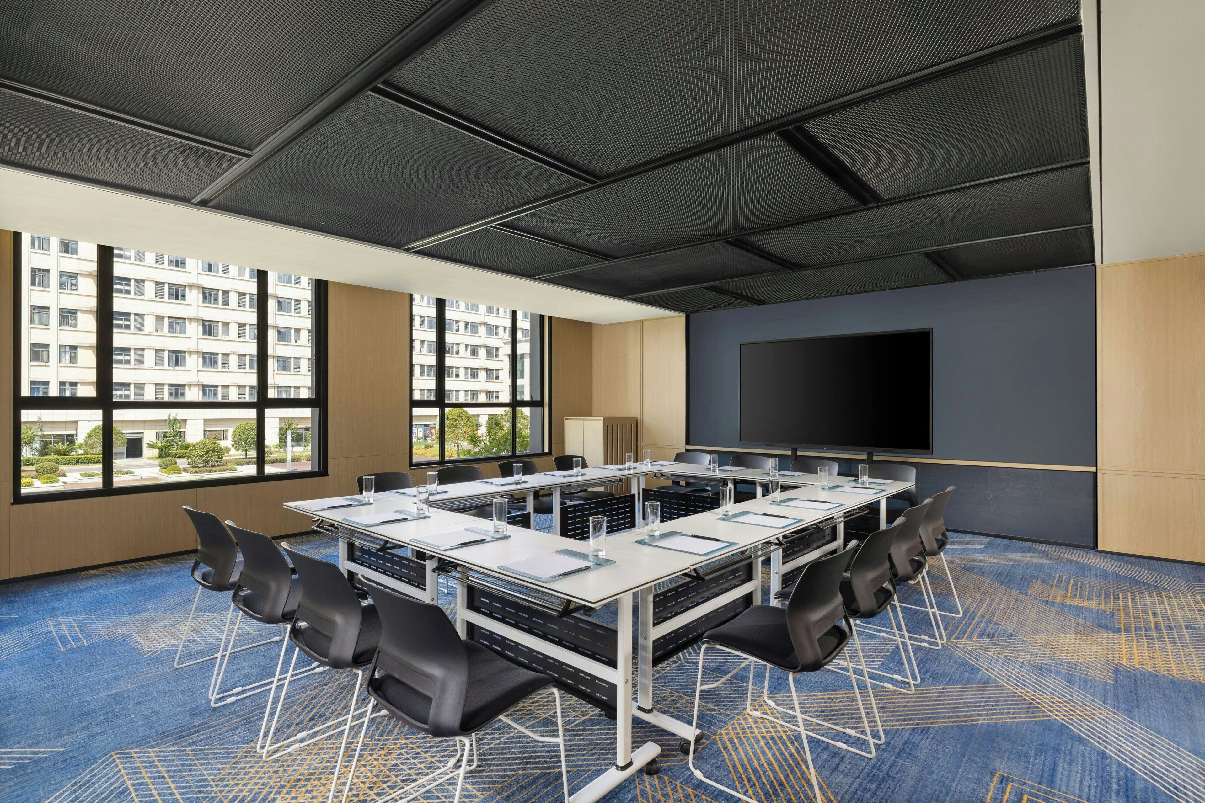 A conference room with a large table, chairs, a projector screen, and large windows with a view of buildings.