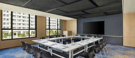 A conference room with a large table, chairs, a projector screen, and large windows with a view of buildings.