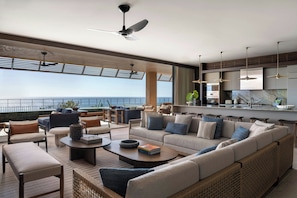 A modern living room with a sofa, coffee table, and chairs, a kitchen area with pendant lights, and a view of the sea.
