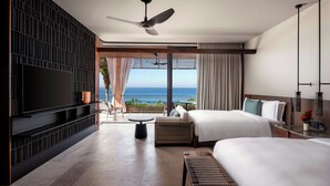 Quarto de hotel com uma televisão grande, ventoinha no teto, cama, mesa de cabeceira, uma mesinha e vista para o mar.