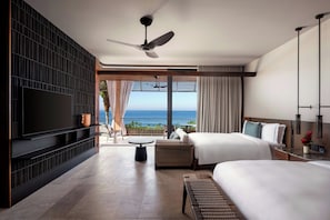 A hotel room with a large TV, a ceiling fan, a bed, a bedside table, a small table, and a view of the ocean.