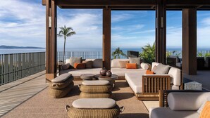 Uma área de estar coberta ao ar livre com vista para o oceano, com mobiliário de vime e uma mesa de pedra.